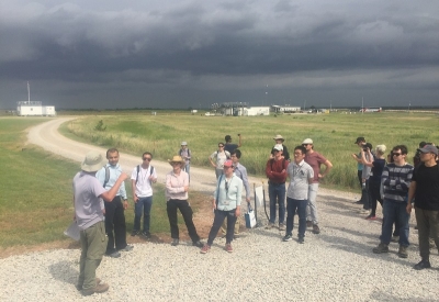 On a stormy-looking day, a person gestures while talking to a group of people along a road in the middle of ARM's Southern Great Plains Central Facility. An Aerosol Observing System and offices and labs near the main entrance are visible in the background. 