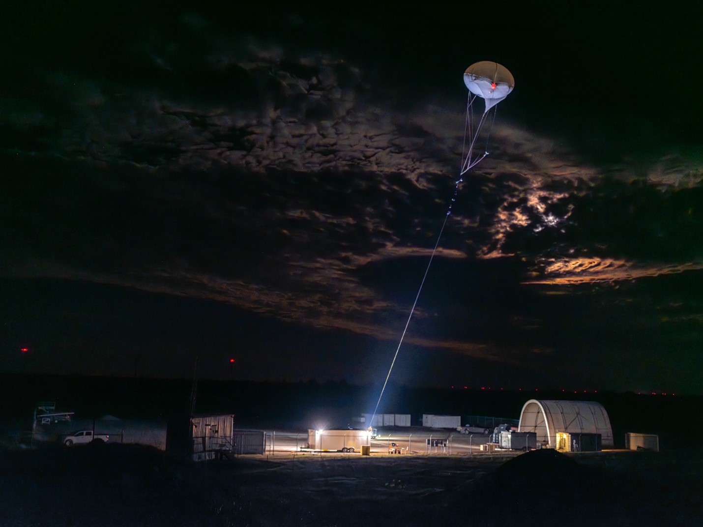 A tethered balloon system is backlit by the moon in a dark, cloudy early morning sky. Lights shine from the ground at the base of the tether. 