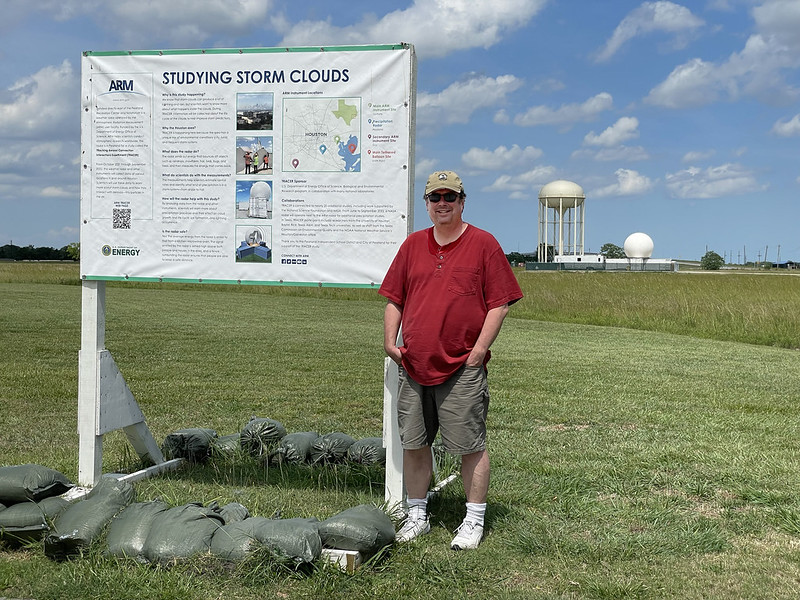 Wearing a red T-shirt and brown cargo shorts, Michael Jensen stands next to a poster titled "Studying Storm Clouds" in front of a water tower and ARM radar.