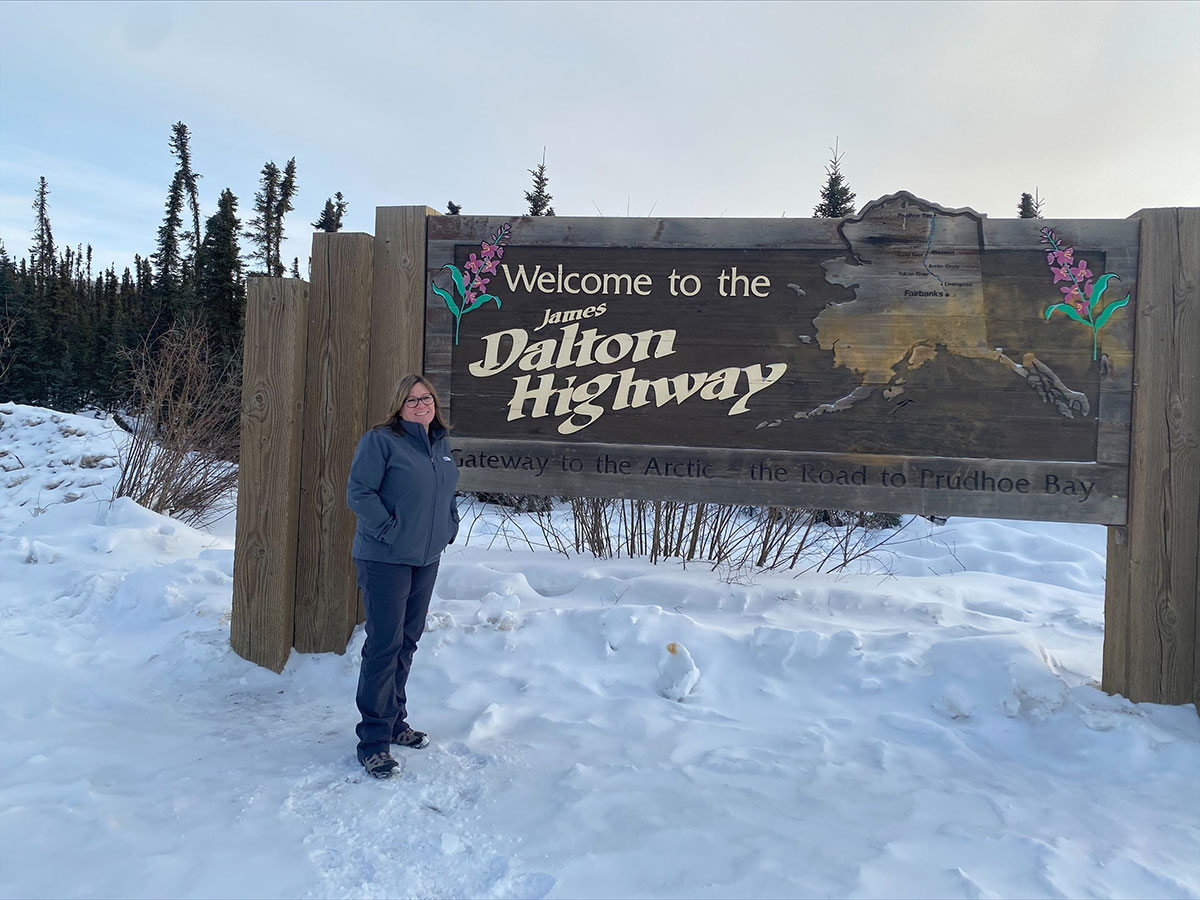Valerie Sparks stands next to a sign saying "Welcome to the James Dalton Highway: Gateway to the Arctic - the Road to Prudhoe Bay"