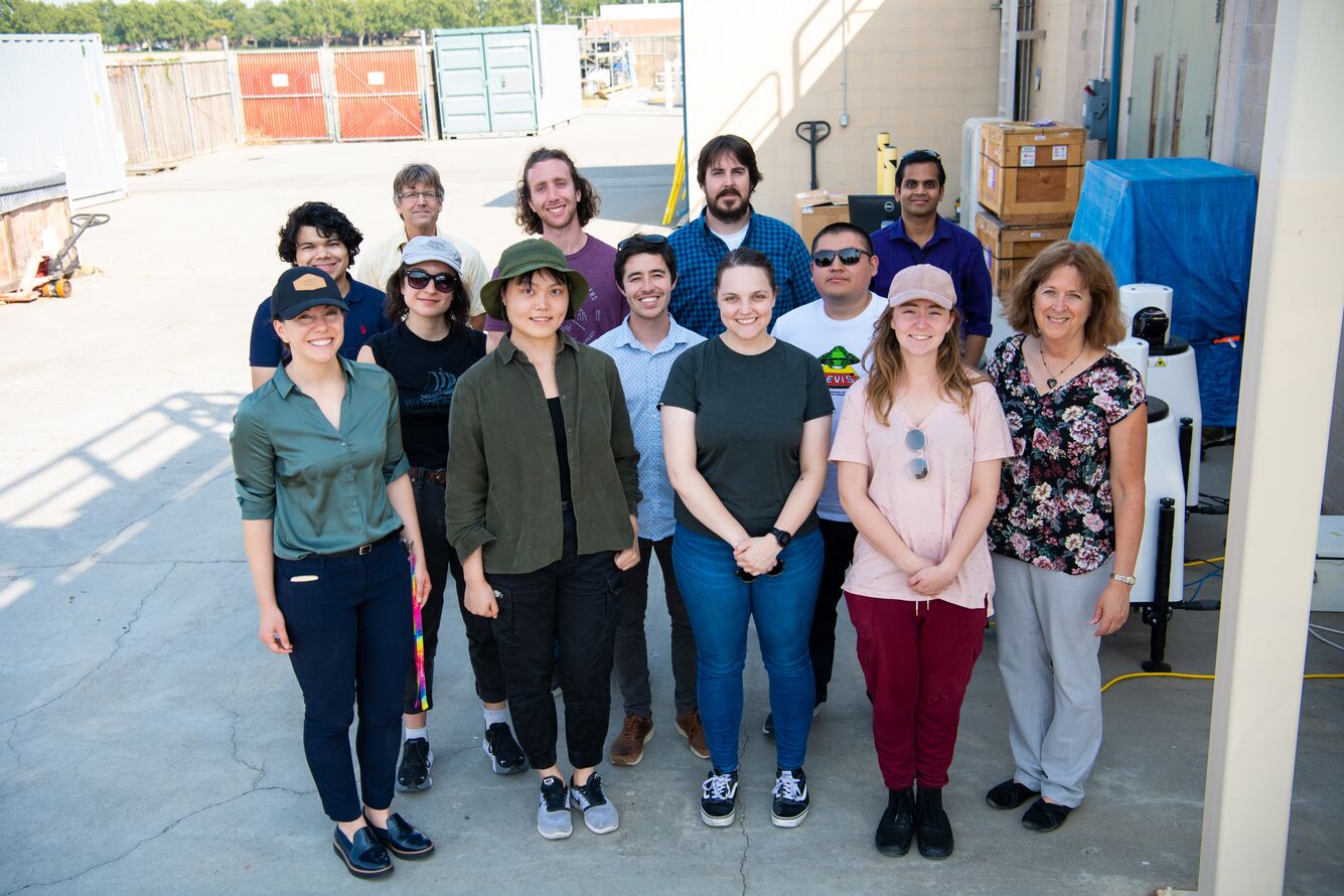 Thirteen people stand together to pose for a picture near a building with boxes stacked along its outside.