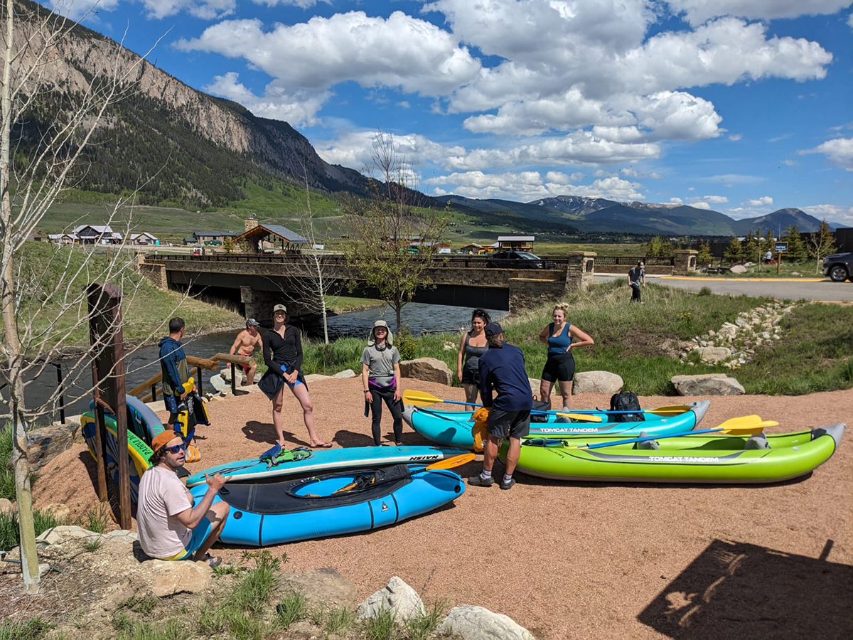 Crew members stand along the river with their inflatable kayaks on the ground.