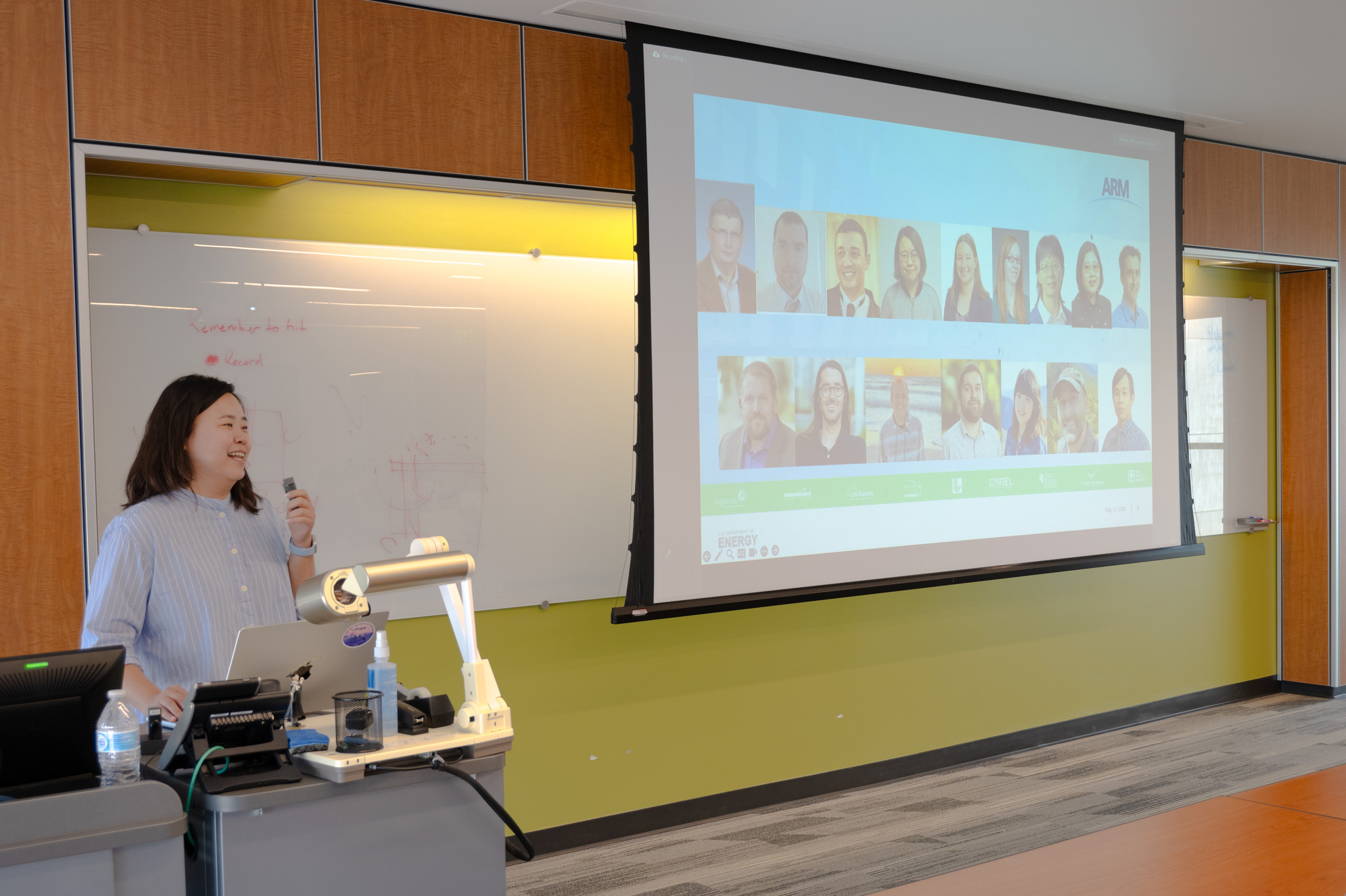 Ya-Chien Feng stands behind a laptop at a podium with a giant screen next to her showing 16 headshots of ARM radar team members.