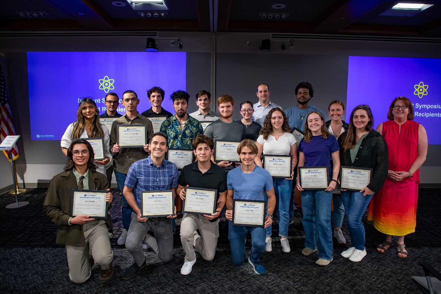 Nineteen people pose together in front of two large screens. Four are kneeling in the front row, and the rest are standing behind them. Most of the people are holding award certificates. 