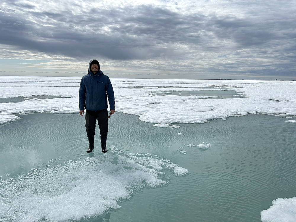 Wearing a dark hooded sweatshirt, pants, and galoshes on a cloudy day, Zac Espinosa stands in melt water on sea ice with patchy snow cover on top.