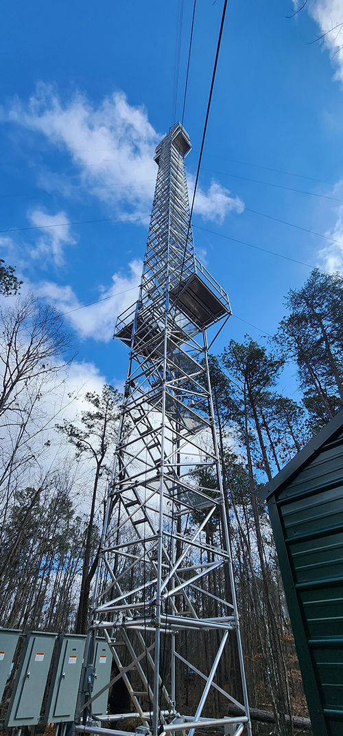 A shot looking up at the 140-foot tower with clouds floating above and tree leaves and branches seen in the bottom corners of the photo