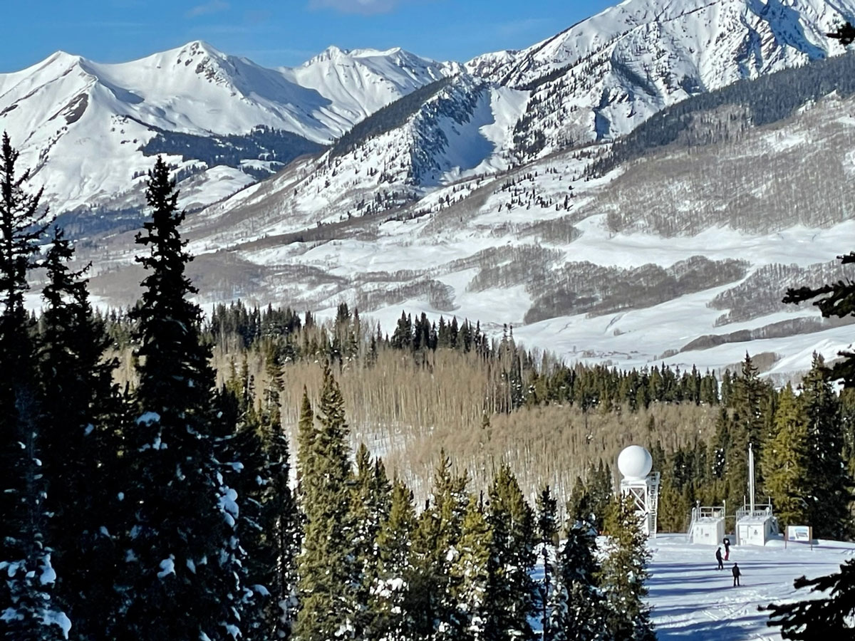 Trees surround the Aerosol Observing System and X-band radar, with snow-covered mountains in the distance.