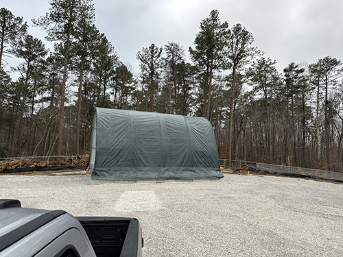 A tethered balloon system hangar sits in a gravel area surrounded by trees. The roof and bed of a gray pickup truck are visible in the bottom left corner of the photo.