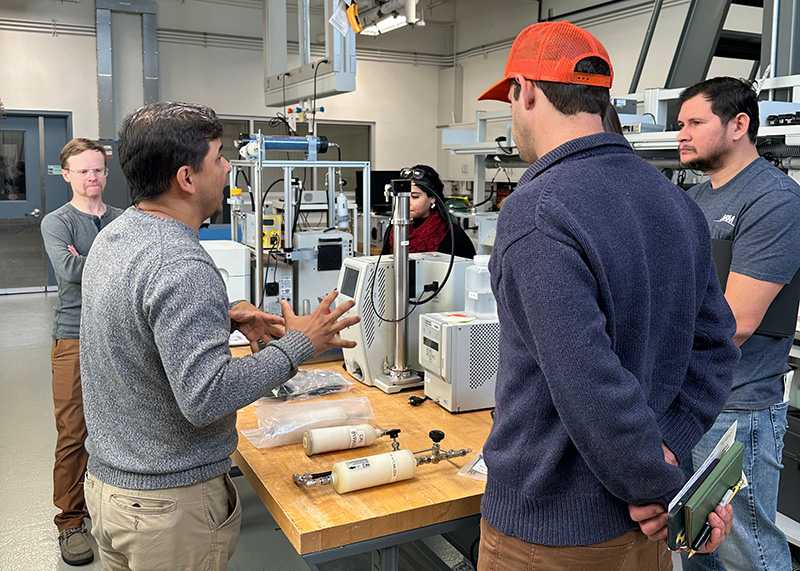 Site operators stand around a work table listening to their instructor's lesson on the scanning mobility particle sizer calibration system.