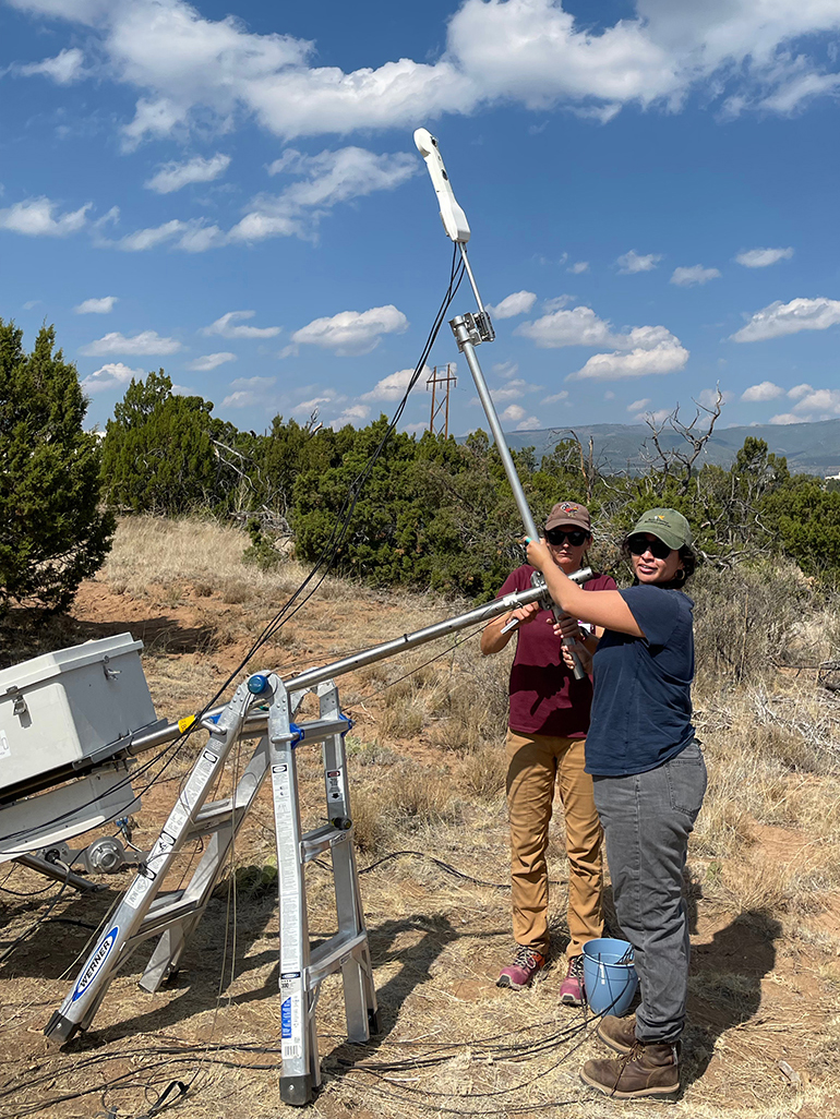 Standing outside at the LANL instrument testing site, Luz Salinas looks at the camera while gripping part of the eddy correlation flux system. Julie Donohue works to her right.