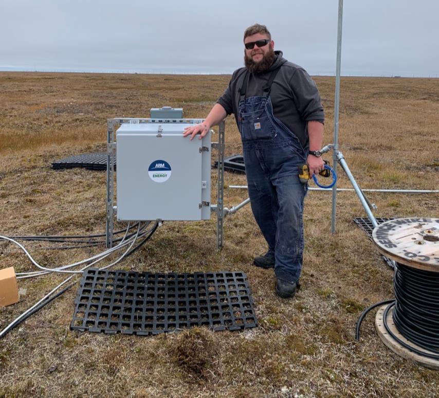 Justin LaPierre stands on the tundra at the E12 site next to a gray box with an ARM/DOE sticker on it. The sky is overcast.