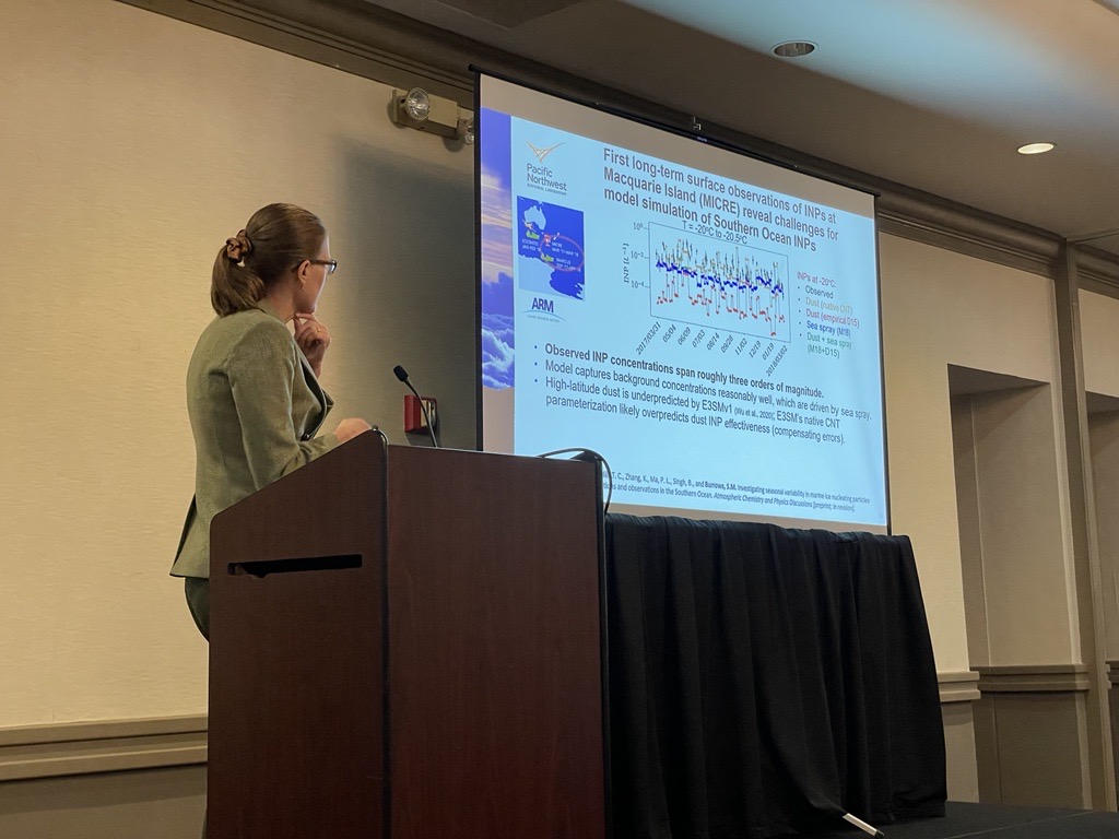 Standing at a podium, Susannah Burrows is turned with her back to the camera while looking at her slide titled "First long-term surface observations of INPs at Macquarie Island (MICRE) reveal challenges for model simulation of Southern Ocean INPs."