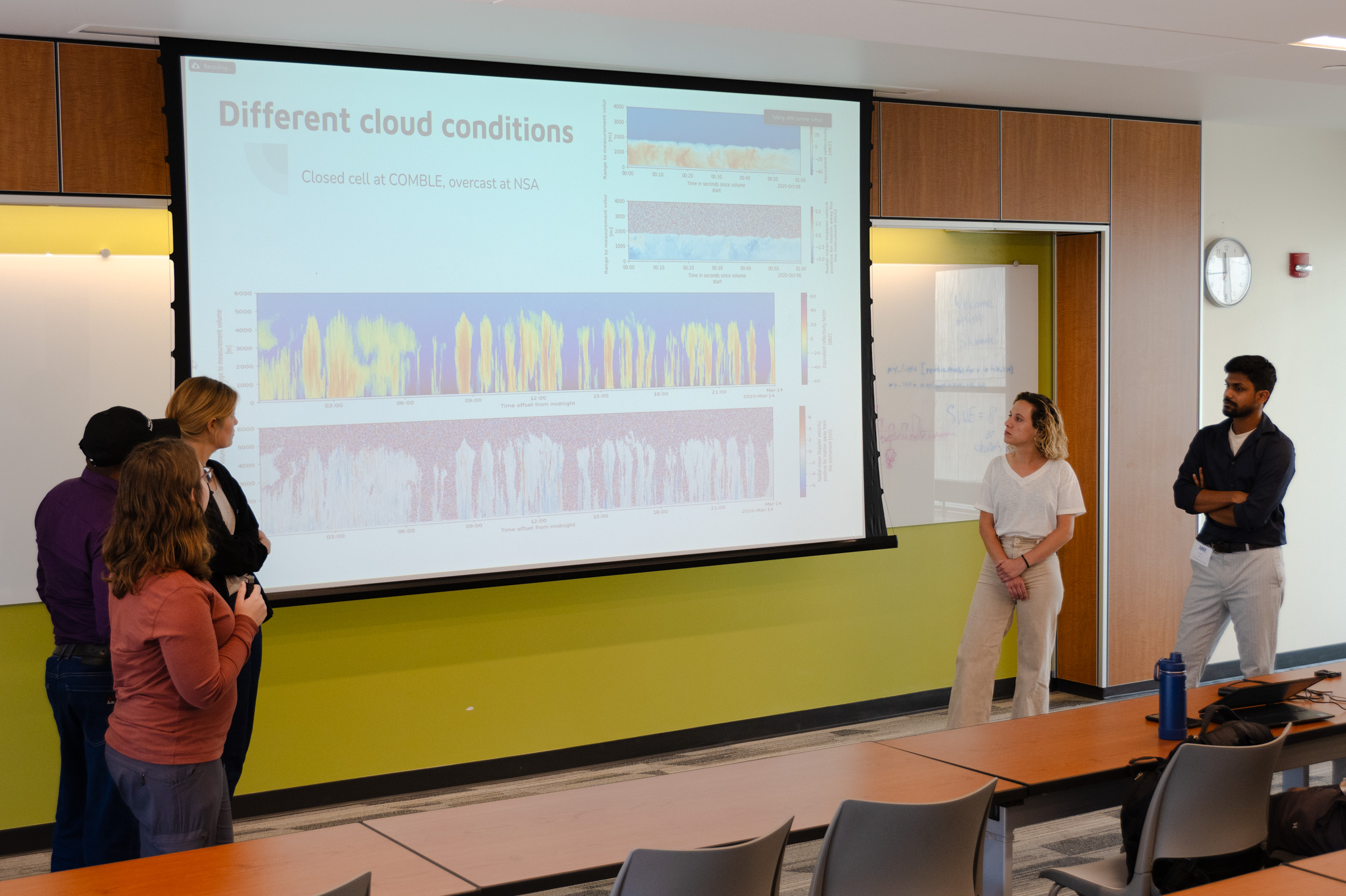 Three members stand on the left side of a giant screen, and two members stand to the right. The screen shows data on different cloud conditions, including closed cell at COMBLE and overcast at the NSA.