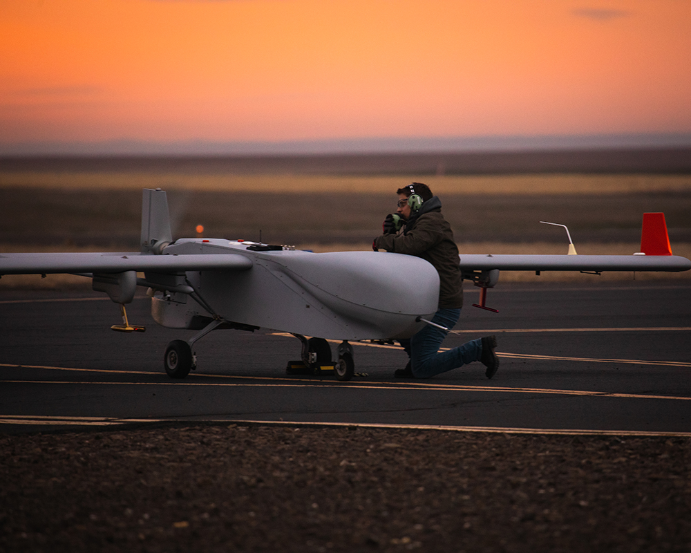 Technician Jonathan Stratman wears a headset, brown jacket, blue jeans, and boots while kneeling next to the ArcticShark uncrewed aerial system and talking into a radio.